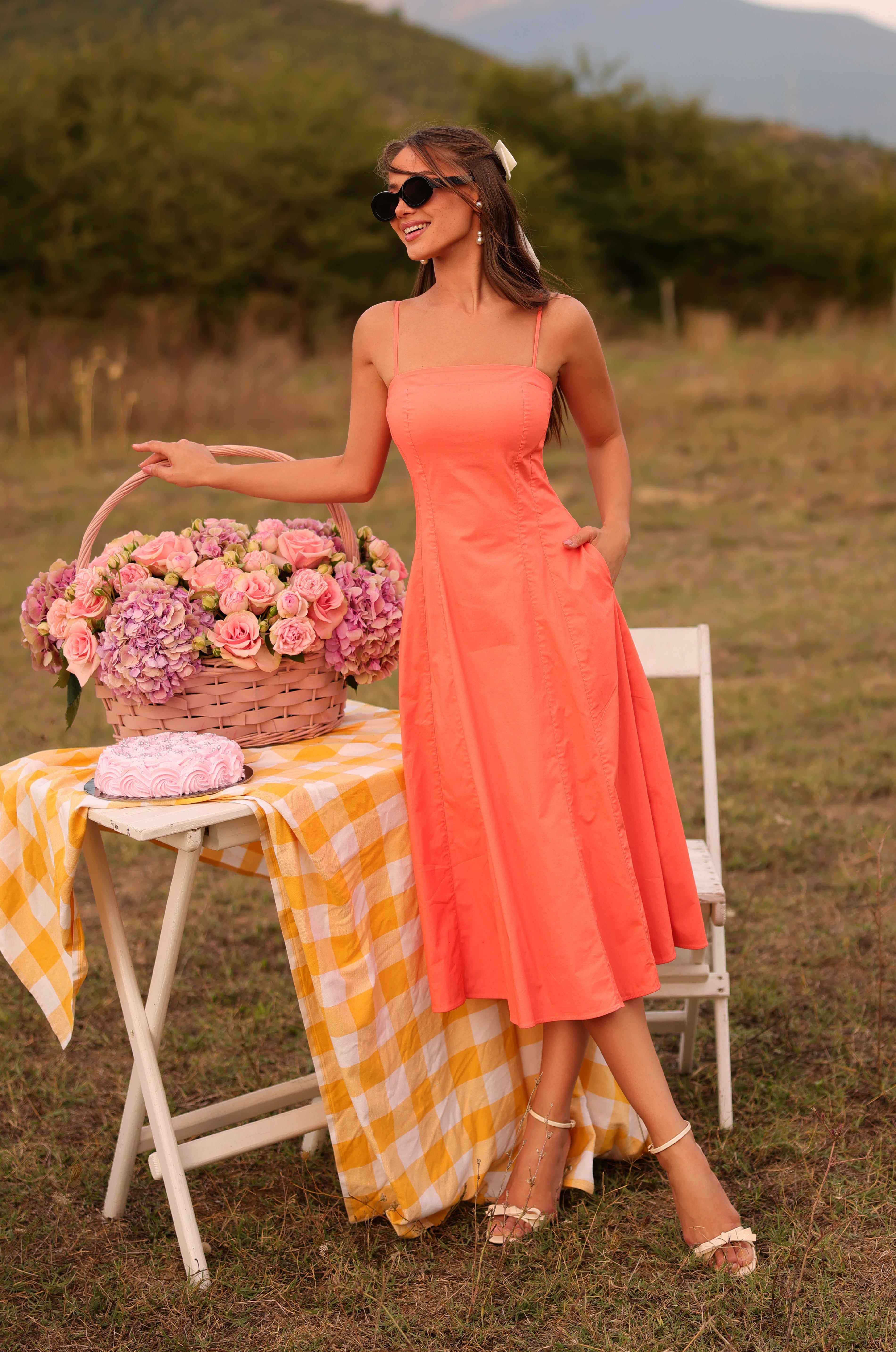 A woman wearing the Papaya Midi Casual Summer Dress in Solid Papaya Punch stands outdoors by a table with a basket of pink flowers, a yellow checkered cloth, and a folded scarf. She smiles and wears sunglasses.